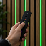Hand holding a black RGB remote control in front of green illuminated timber wall slats